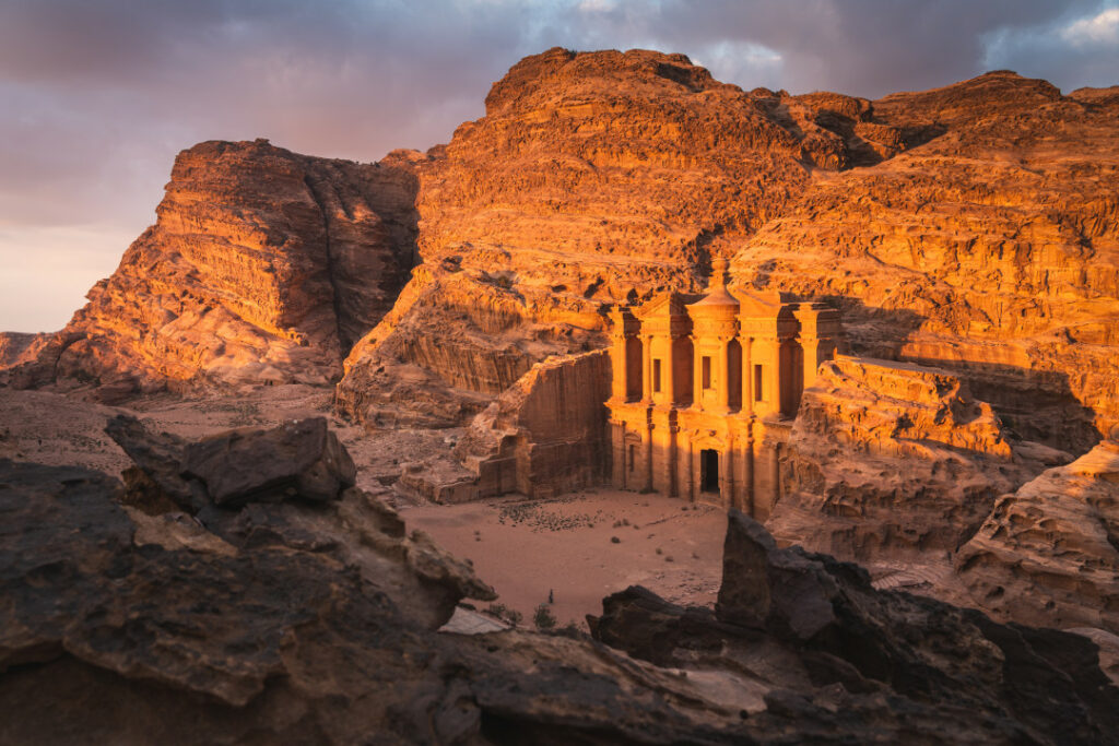 The image showcases the breathtaking view of The Monastery, also known as Ad Deir, bathed in the warm glow of the setting sun. The ancient Nabatean city of Petra comes to life in this stunning sunset scene, revealing the majesty and grandeur of its well-preserved ruins.